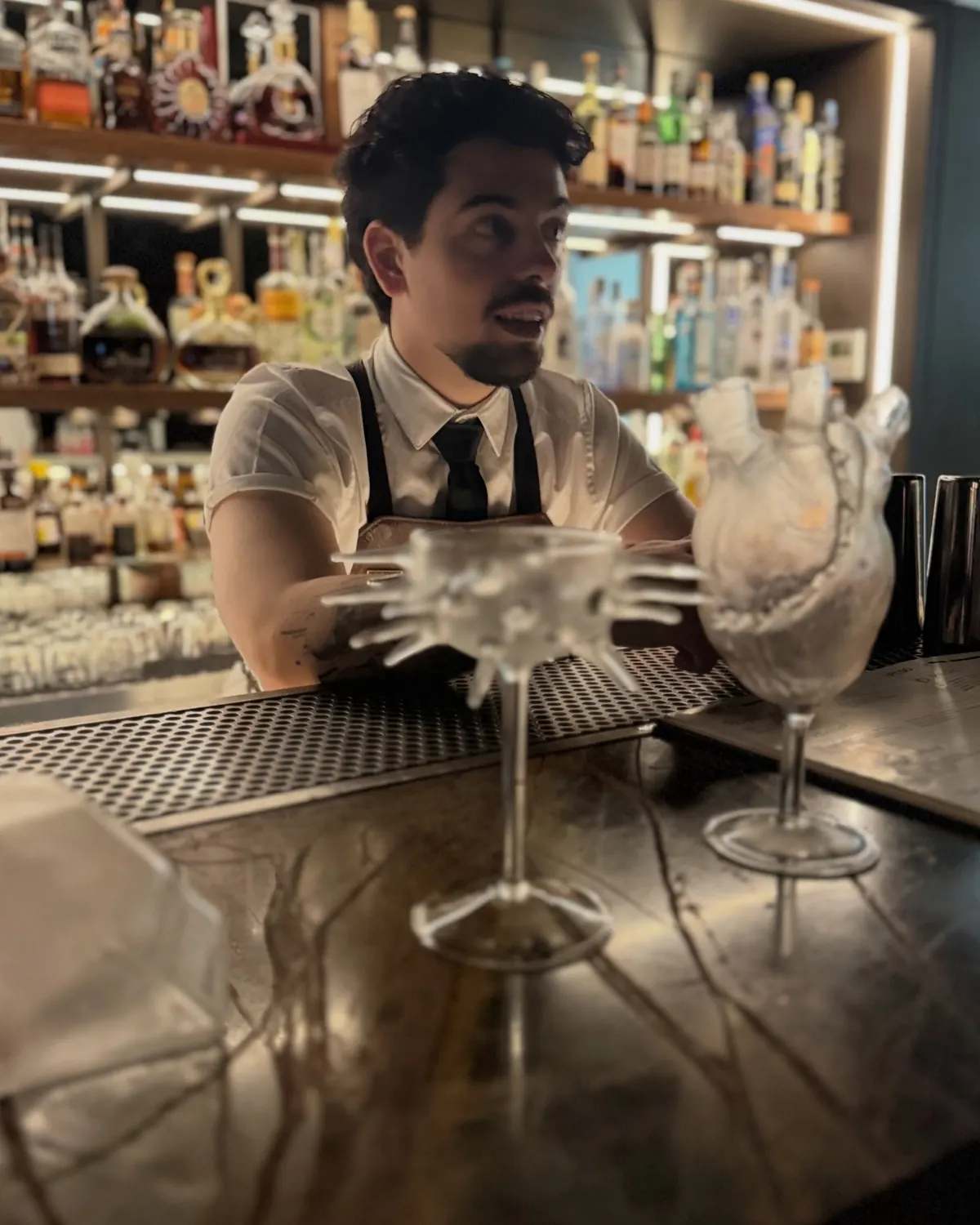 A smiling bartender leans on a polished bar, bottles and glassware behind him.